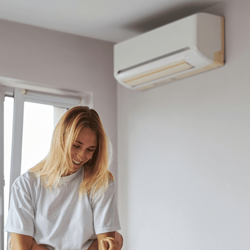 A smiling young woman in a white t-shirt enjoying the cool air from a wall-mounted air conditioner in a light-colored room.